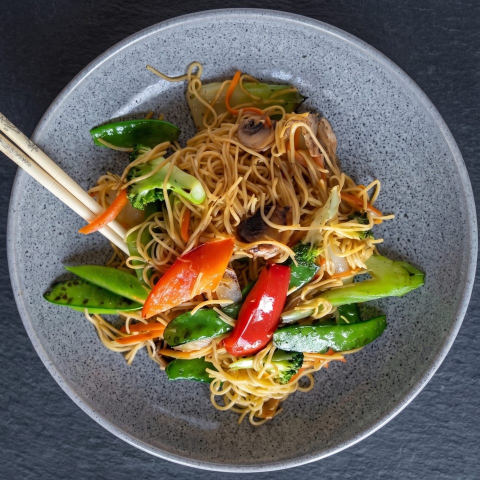 A grey bowl of vegetable lo mein with chopsticks, featuring noodles, broccoli, snap peas, and peppers.