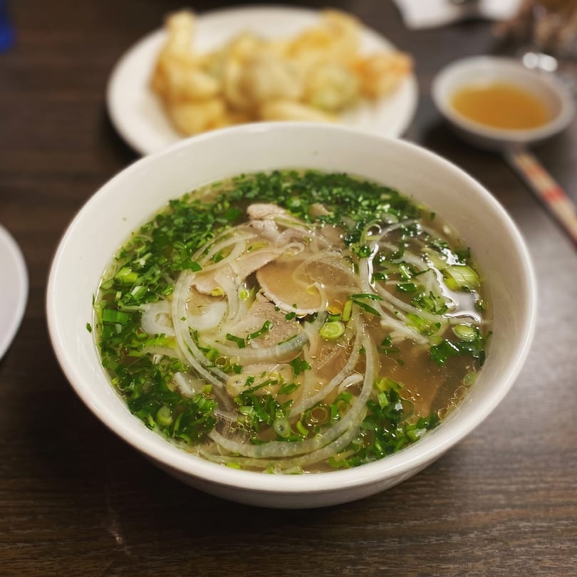 A white bowl of pho with beef, noodles, onions, and green herbs on a dark wooden table.