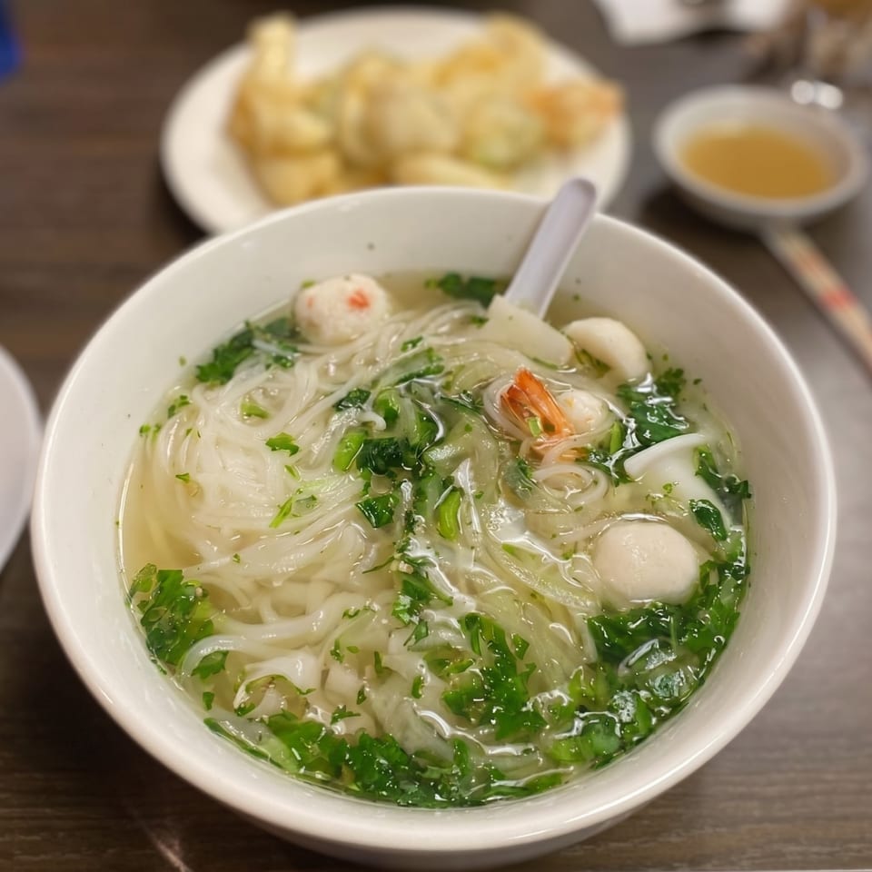 A bowl of pho with noodles, shrimp, fish balls, and green herbs, with appetizers in the background.
