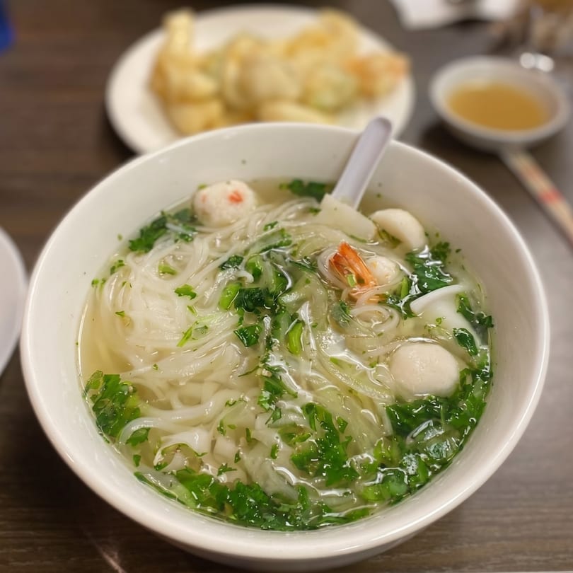 A bowl of pho with noodles, shrimp, fish balls, and green herbs, with appetizers in the background.