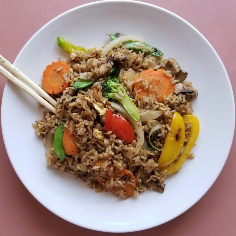 Vegetable fried rice with carrots, bell peppers, broccoli, and snap peas on a white plate with chopsticks.