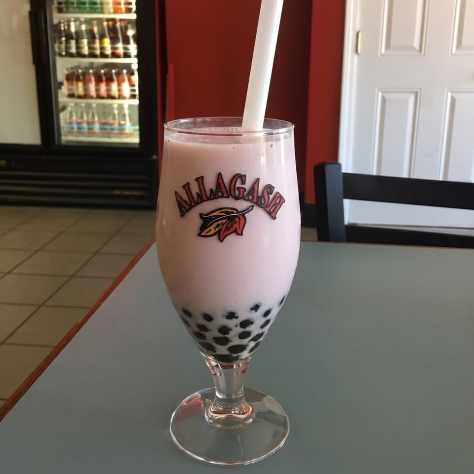 Pink boba tea in an "Allagash" branded glass with a white straw on a table.