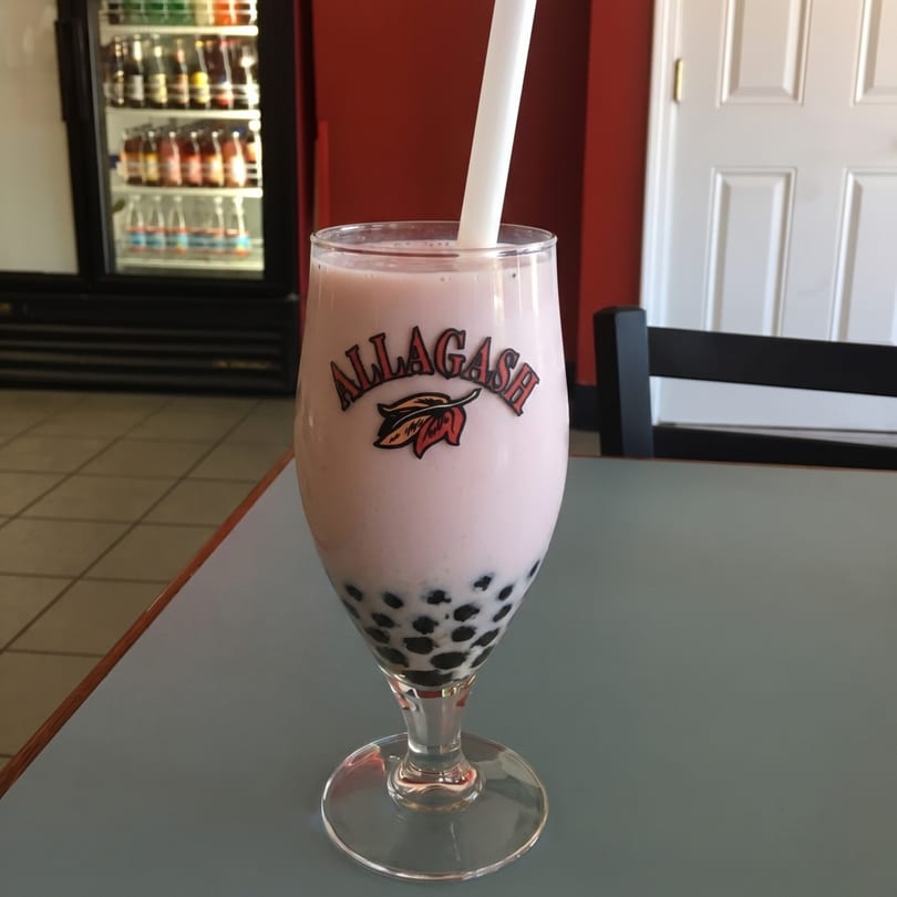 Pink boba tea in an "Allagash" branded glass with a white straw on a table.