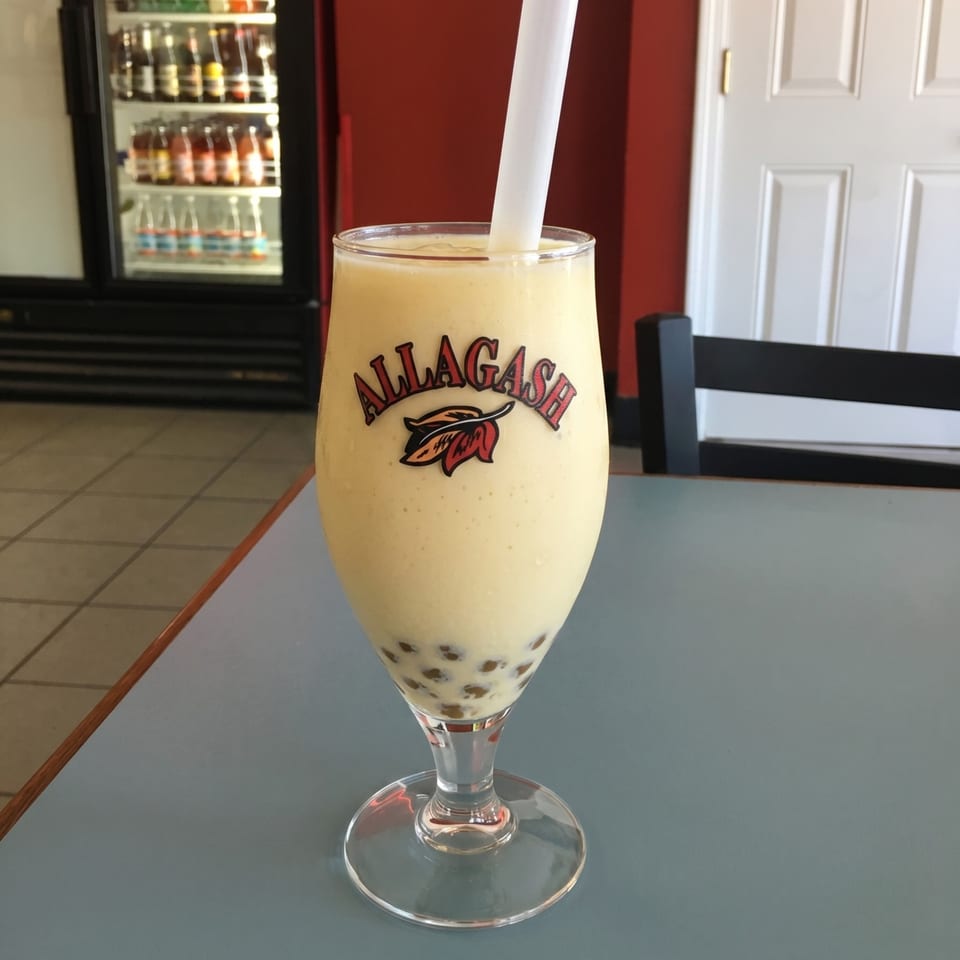 Allagash branded glass with a light yellow boba drink and white straw on a blue table.